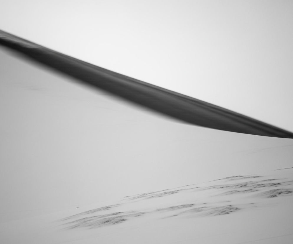 Minimalistyczną, czarno-białą kompozycję abstrakcyjną. U góry, skośnie od lewego górnego rogu do prawego środka, przebiega szeroki, rozmyty pas w ciemnym odcieniu szarości, niemal czerni, z nieregularnymi, jaśniejszymi smugami wzdłuż jego lewej krawędzi, sugerującymi ruch lub teksturę. Pozostała część obrazu to jasna, jednolita płaszczyzna w odcieniach bieli i jasnej szarości, z subtelnymi, ciemniejszymi plamami i liniami w dolnej części, tworzącymi wrażenie nierównej powierzchni lub cieni. Całość tworzy dynamiczny, choć monochromatyczny pejzaż, pozbawiony ostrych detali.