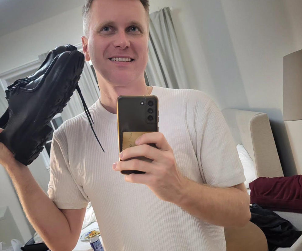 Uśmiechnięty Artur Gotz robi lustrzane selfie, trzymając w górze czarny but. O jego przygodzie w Chicago czytaj na SE USA.