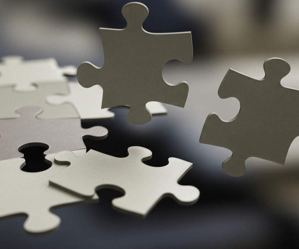 Dwa pojedyncze, szare elementy puzzli unoszą się po prawej stronie kadru, nieco pod kątem, z delikatnym, jasnym światłem odbijającym się od ich górnych krawędzi. Po lewej stronie leży ułożonych kilka innych elementów puzzli w odcieniach szarości i beżu, tworząc częściowo spójną płaszczyznę. Całość spoczywa na ciemnej, błyszczącej powierzchni, która odbija światło z góry. Tło jest rozmyte, w neutralnych, jasnoszarych tonach.