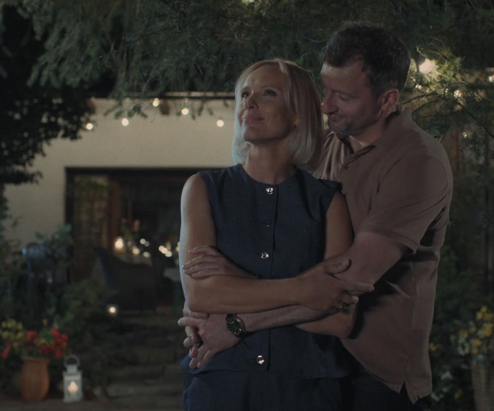 Para, Dorota i Bartek z serialu M jak miłość, wtulona w siebie w ogrodzie siedliska w Grabinie, w tle rozświetlone lampkami dekoracyjnymi. Kobieta ma blond włosy i ciemną sukienkę, a mężczyzna brązową koszulkę. Romantyczny wieczór w Super Seriale.