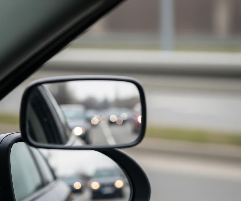 Główne elementy to lusterko wsteczne samochodu i jego odbicie, ukazujące ruch uliczny za pojazdem. Lusterko, obramowane czarną ramką, znajduje się w prawym środkowym kadrze, z fragmentem ciemnego słupka samochodu po lewej stronie. W odbiciu lusterka widoczne są rozmyte światła samochodów na drodze, tworzące jasne, okrągłe punkty, a także fragment białej linii rozdzielającej pasy ruchu. Tło, widoczne za lusterkiem i w jego odbiciu, jest rozmyte, przedstawiając asfaltową drogę, zielony pas pobocza oraz szary mur lub barierkę.