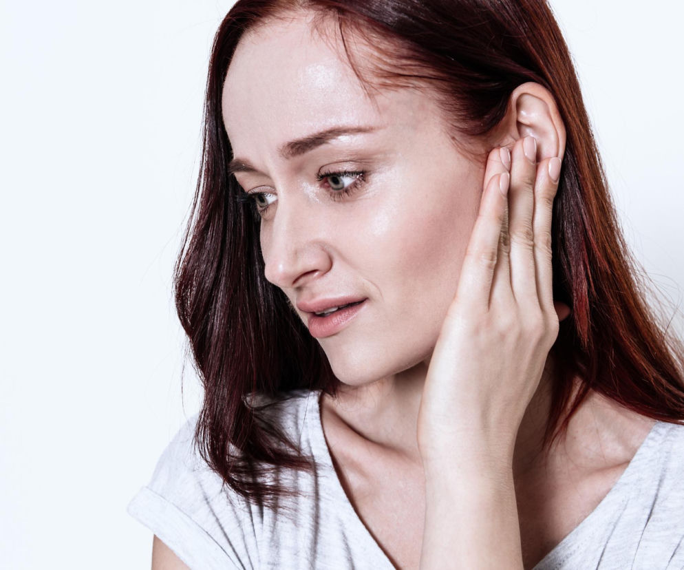 Zaniepokojona kobieta z rudymi włosami trzymająca dłoń przy uchu, symbolizująca dolegliwości lub gorące uszy. Na Poradnik Zdrowie znajdziesz informacje o tym, dlaczego uszy stają się czerwone i gorące, oraz jak odróżnić te objawy od nadciśnienia.