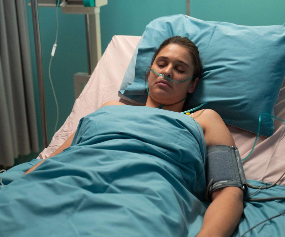 Aktorka Anna Szymańczyk jako Iga Kaczorowska leży na szpitalnym łóżku pod niebieskim kocem, z zamkniętymi oczami i rurką tlenową w nosie, z aparatem do mierzenia ciśnienia na ramieniu. O jej walce o życie po wykryciu tętniaka mózgu i wpływie narkotyków, czytaj na Super Seriale.