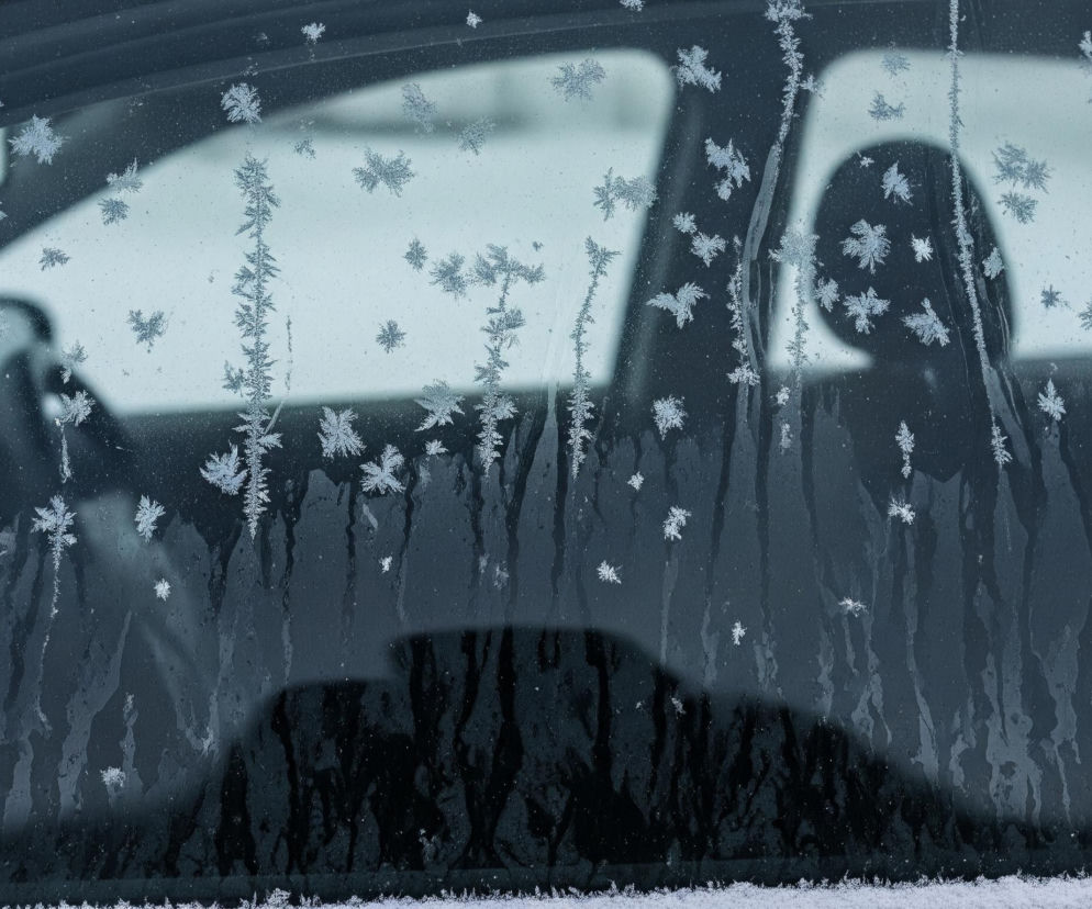 Widoczna jest boczna szyba samochodu pokryta szronem w postaci drobnych kryształków lodu. Szyba jest ciemna, niemal czarna na dole, przechodząc w jaśniejsze odcienie szarości i błękitu ku górze, gdzie widać rozmyte odbicia jasnego otoczenia. Na całej powierzchni szyby rozmieszczone są struktury szronu, przypominające listki paproci lub gwiazdki śniegu, o zróżnicowanej gęstości i rozmiarze. Prawa krawędź szyby i fragmenty lewej krawędzi są obramowane czarną ramą, a wzdłuż prawej ramy tworzy się pionowa, gęsta kolumna kryształków lodu. W dolnej części obrazu, pod szybą, rozciąga się pas białego, puszystego śniegu.