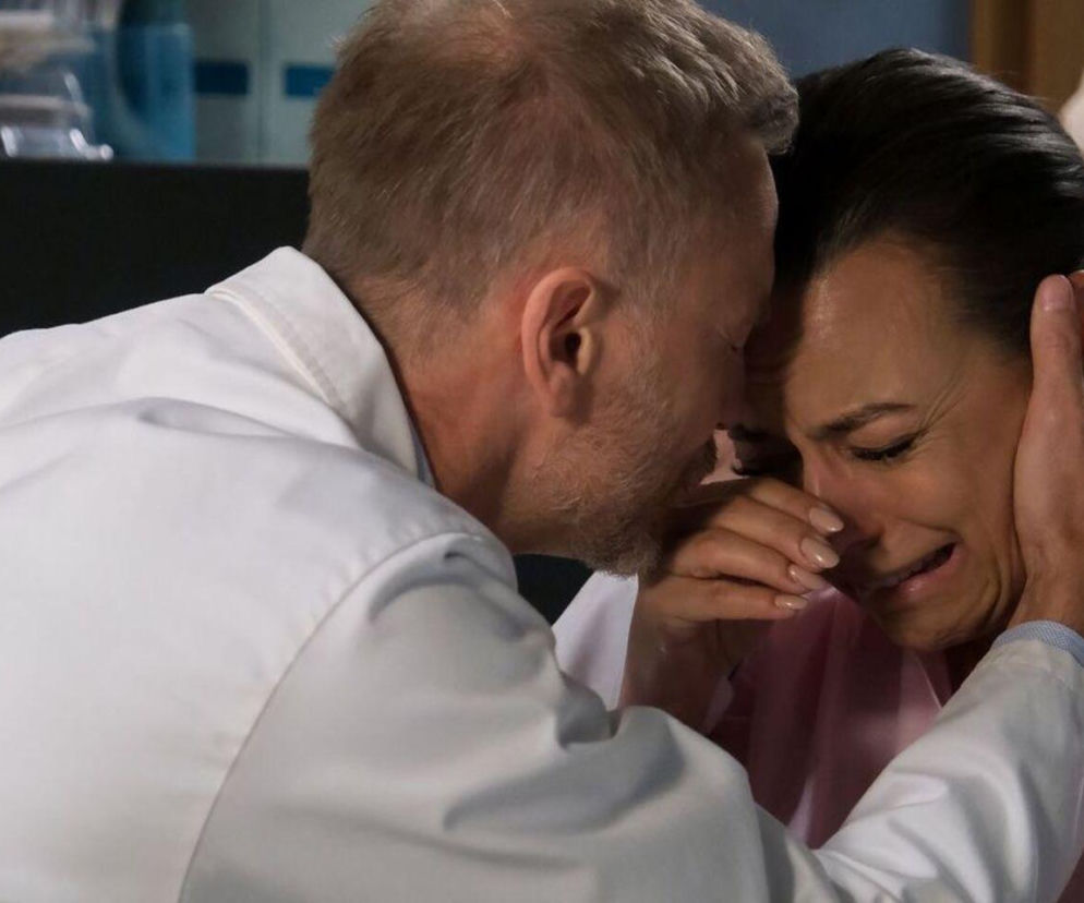 Andrzej Budzyński (Krystian Wieczorek) w białym lekarskim fartuchu pociesza zapłakaną Magdę (Anna Mucha), która jest aresztowana. Mężczyzna trzyma jej twarz w dłoniach, a ona ociera łzy. Scena pełna emocji z serialu M jak miłość. Więcej o tym na stronie Super Seriale.