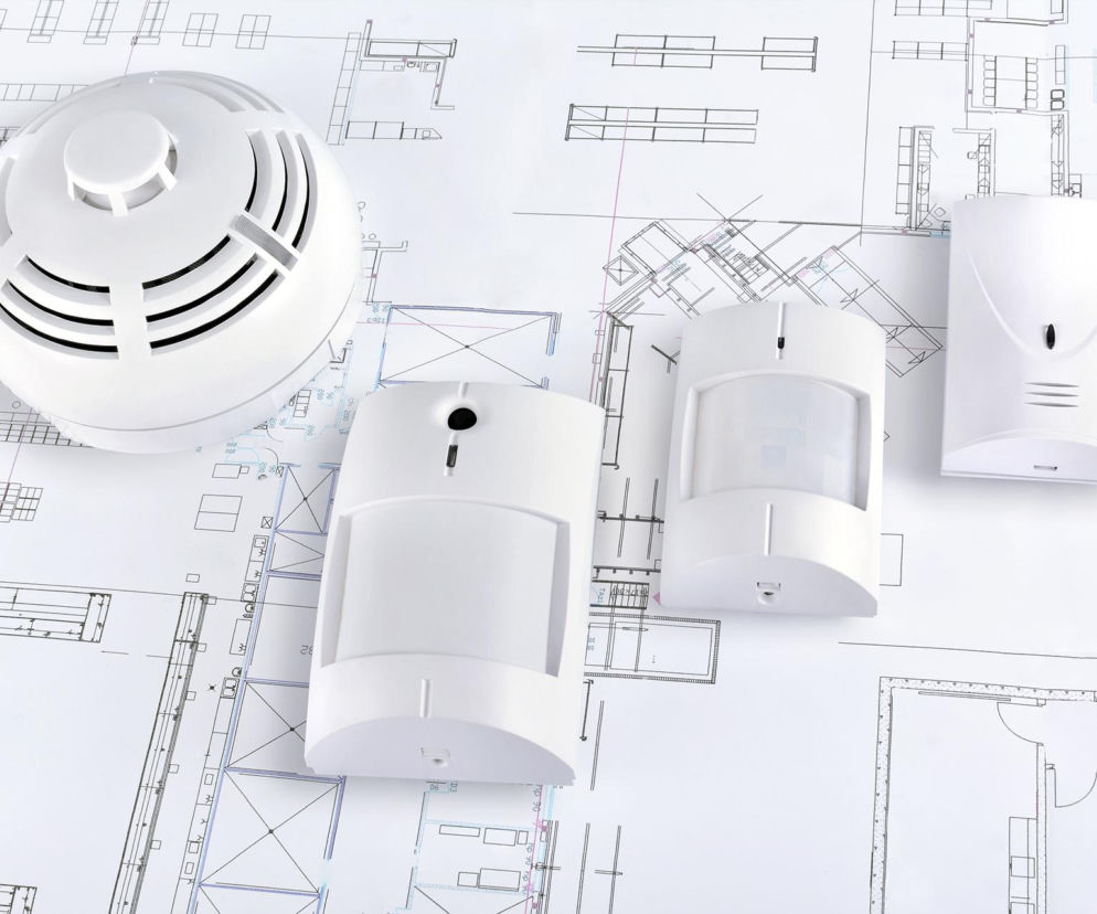 Białe czujki alarmowe leżą na projekcie architektonicznym domu. O planowaniu systemów bezpieczeństwa pisze Muratordom.