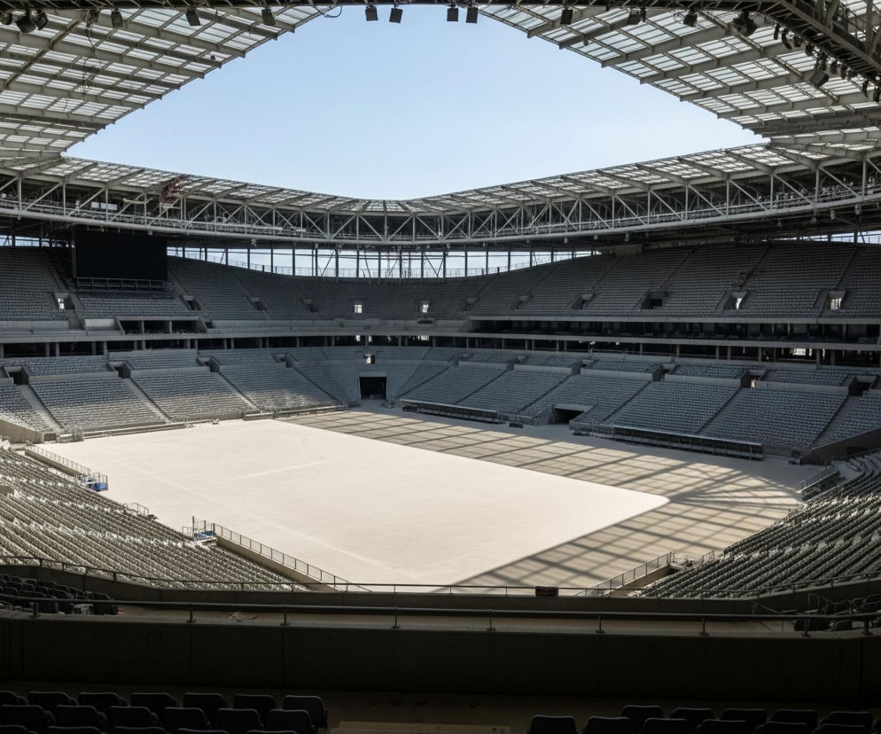 Pusty, nowoczesny stadion sportowy z licznymi rzędami szarych siedzeń wypełnia kadr, widziany z góry. Centralnie położone boisko jest pokryte jasnym, beżowym materiałem, a na jego powierzchni widoczne są cienie konstrukcji dachu. Składany dach, częściowo przezroczysty, składa się z metalowych belek i paneli, otaczając otwarty, błękitny fragment nieba. Na górze widać rzędy reflektorów przymocowanych do konstrukcji.