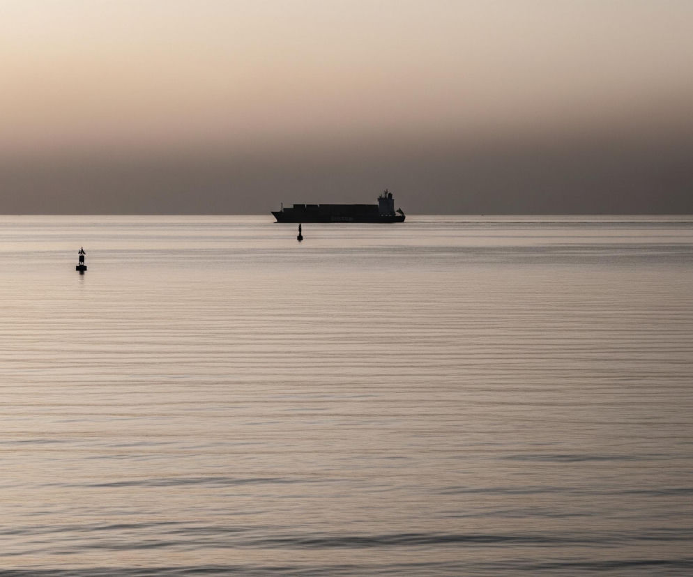 Kontenerowiec płynie na spokojnym, lekko pofałdowanym morzu, w oddali, pod horyzontem. Przed nim, bliżej centrum kadru, unosi się czarno-biała boja, a dalej na prawo widać mały, ciemny obiekt. Niebo nad morzem rozjaśnia się od góry, przechodząc od delikatnego brązu do jasnego beżu tuż nad horyzontem, a ta sama ciepła paleta barw odbija się w wodzie, tworząc jednolity, spokojny nastrój.