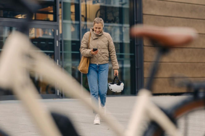 Uśmiechnięta kobieta w beżowej kurtce i dżinsach, z torebką na ramieniu, idzie ulicą, patrząc w smartfon i trzymając kask rowerowy. Obok niej widać zamazany rower, co nawiązuje do ekosystemu Bosch Connected Biking na Super Biznes.