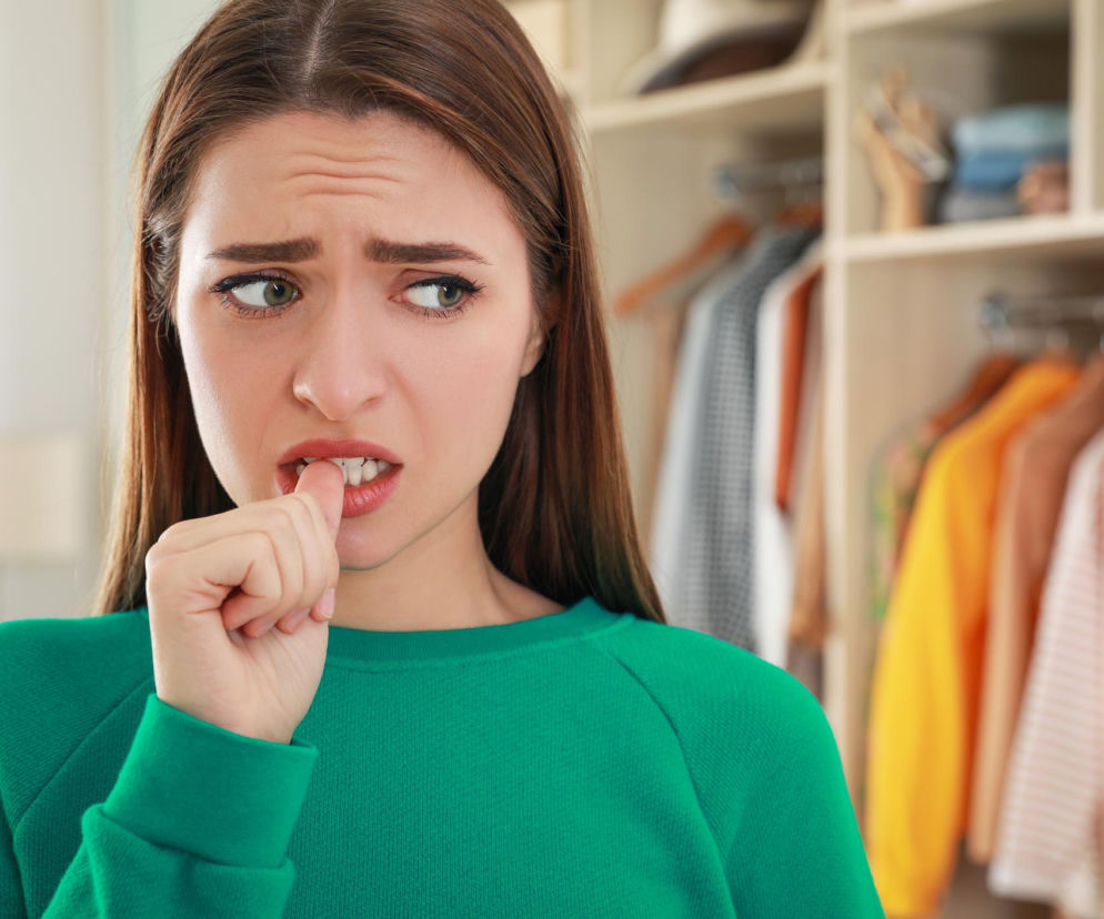 Kobieta zaniepokojona lub zestresowana, z brązowymi włosami i marszczonymi brwiami, obgryzająca paznokcie, ubrana w zieloną bluzę, na tle otwartej szafy z ubraniami. Artykuł Poradnik Zdrowie wyjaśnia, jak poprawić koncentrację i radzić sobie ze stresem.