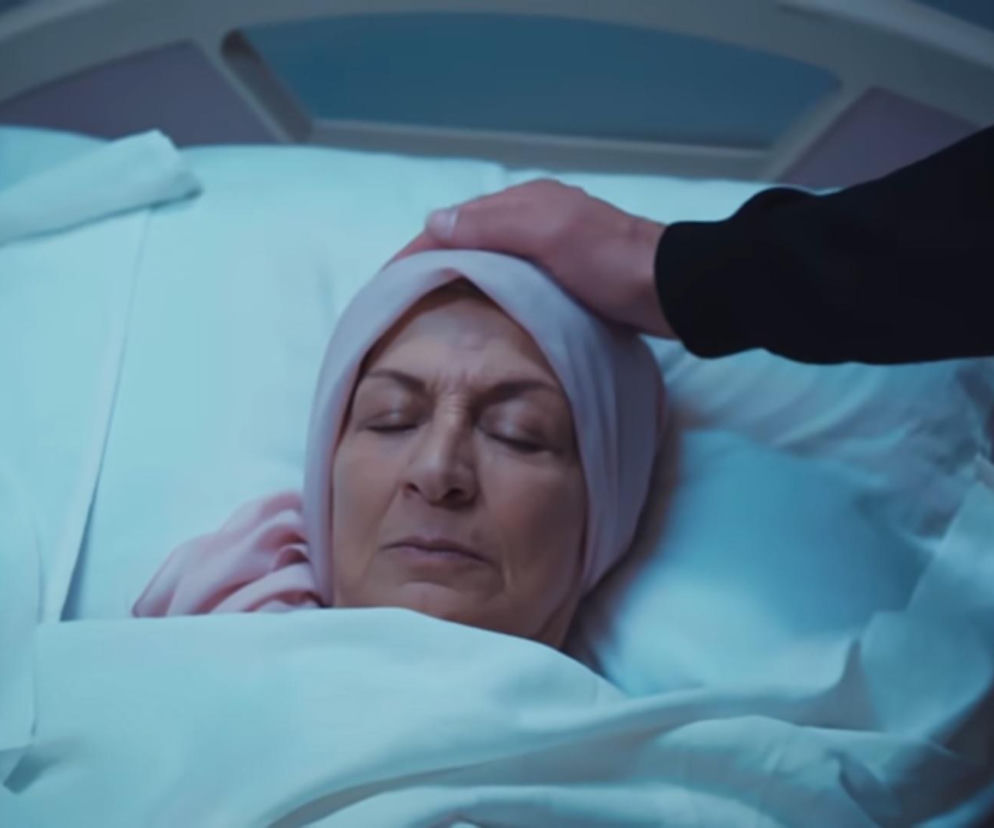 Kobieta w jasnoróżowym czepku leży w szpitalnym łóżku, a męska dłoń delikatnie spoczywa na jej głowie, oddając ostatni hołd Akcy z serialu Dziedzictwo. Zdjęcie to ilustruje dramatyczne pożegnanie z bliską osobą. Więcej o tym odcinku znajdziesz na Super Seriale.