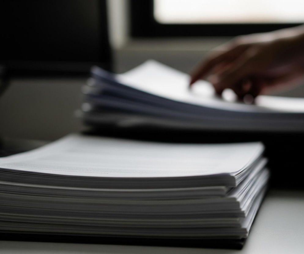 Na pierwszym planie, z lewej strony, widnieje stos białych kartek papieru, ułożony na ciemnej podkładce, leżący na jasnym blacie. Za nim, częściowo niewidoczna, znajduje się czarna klawiatura oraz monitor. W tle, po prawej stronie, widać kolejny, wyższy stos papieru, z którego dłoń w mankiecie ciemnej koszuli i marynarki właśnie wyjmuje pojedynczą kartkę. Z tyłu, po prawej stronie, widoczne jest rozmyte okno wpuszczające światło, kontrastujące z ogólnym, nieco przyciemnionym oświetleniem sceny.