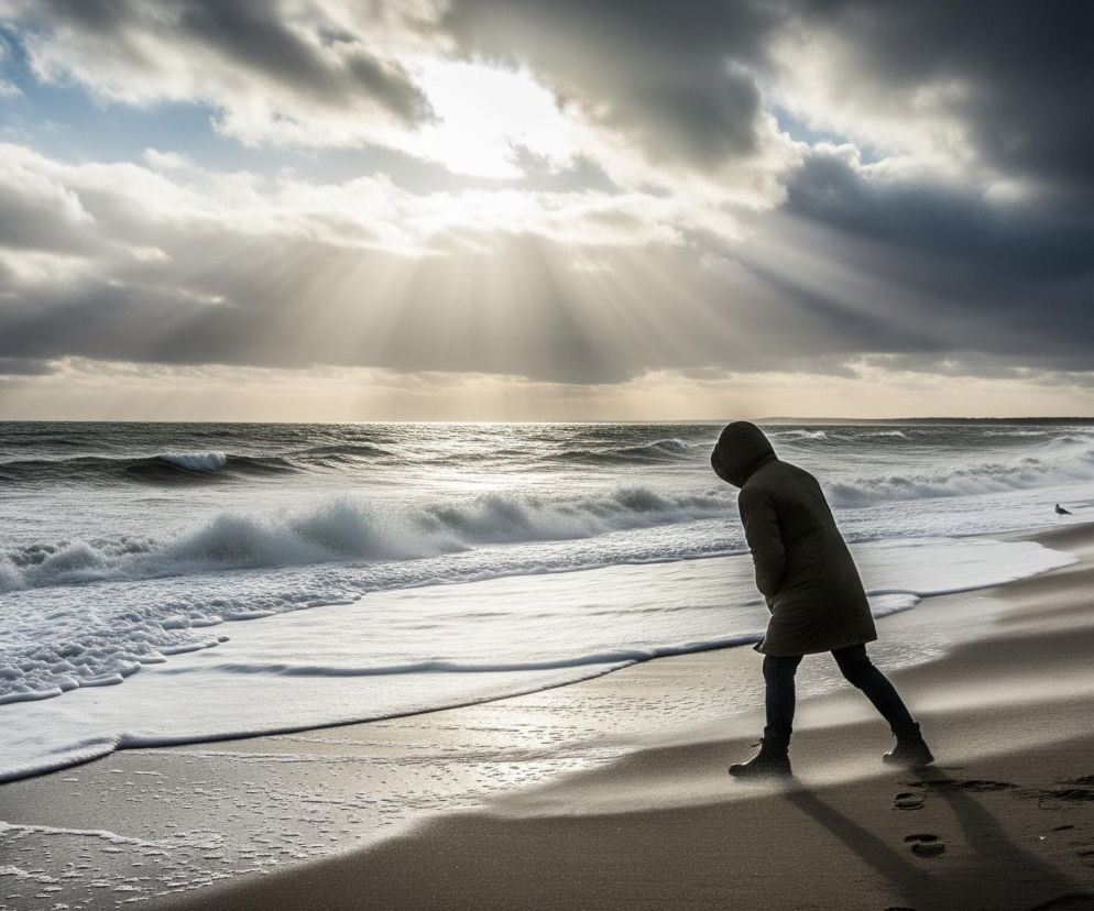 Osobę w ciemnej, długiej kurtce z kapturem, pochyloną, stojącą na piaszczystej plaży. Słońce, częściowo zasłonięte przez ciemne chmury, przebija promieniami od góry, tworząc srebrzyste refleksy na falującej wodzie i jasne pasma na piasku. Brzeg morza pokrywają białe, spienione fale, a po prawej stronie plaży, dalej od postaci, widoczne są pojedyncze mewy.