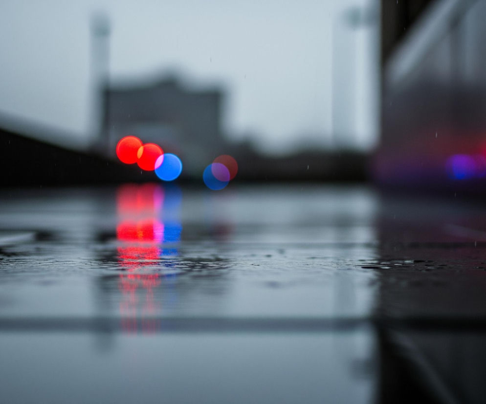 Na pierwszym planie widoczna jest mokra, ciemna powierzchnia, prawdopodobnie asfalt lub bruk, pokryta kroplami deszczu, tworząca odbicia. W głębi obrazu, w centrum kadru, znajdują się trzy wyraźne, rozmyte światła w kształcie kół – dwa czerwone i jedno niebieskie – z lekkimi refleksami poniżej. Dalej w tle, po prawej stronie, widać kolejne, bardziej rozmyte, czerwone i niebieskie punkty świetlne. Całe tło jest mocno nieostre, ukazując szare, zamazane kształty budynków lub konstrukcji, a z góry spadają pojedyncze, ledwo widoczne krople deszczu, tworząc wrażenie deszczowego dnia.
