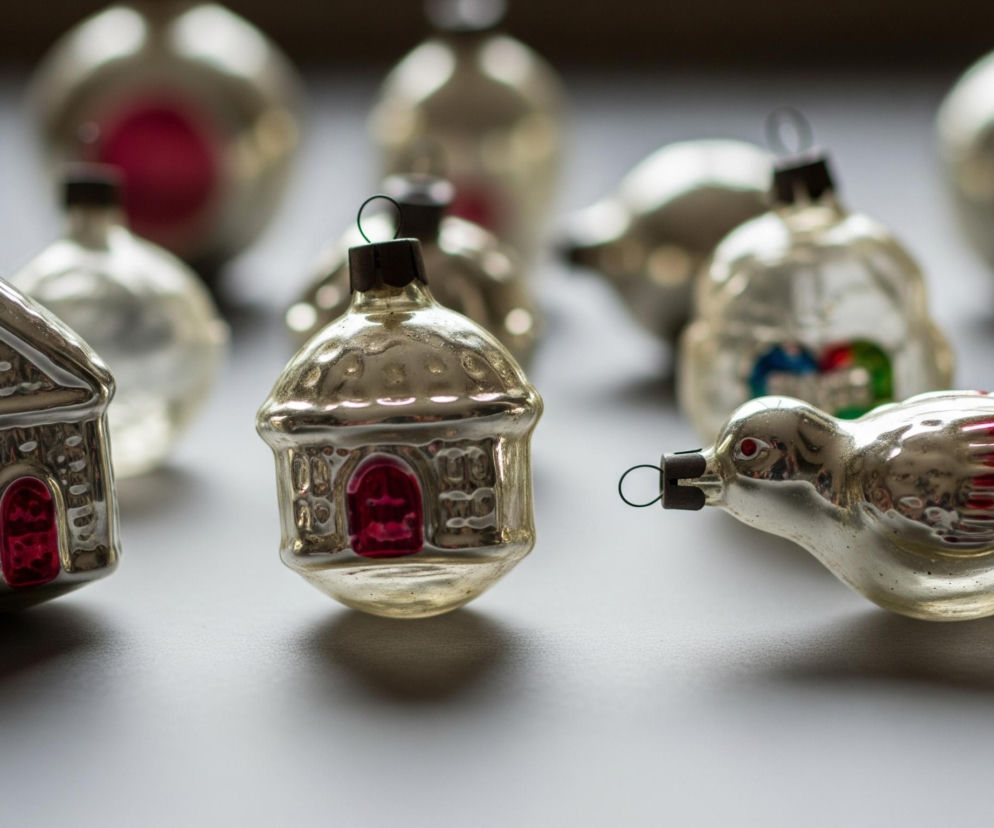 Na szarym blacie leżą błyszczące, szklane ozdoby choinkowe. W pierwszym rzędzie, od lewej, znajduje się srebrny domek z czerwonymi drzwiami i oknami, obok niego stoi drugi, bardziej zaokrąglony, również srebrny domek z czerwoną wnęką. Po prawej stronie leży srebrny ptak z czerwonym okiem i czerwonymi piórami na skrzydłach. W tle, częściowo rozmazane, widoczne są inne błyszczące ozdoby, w tym kilka w kształcie kuli z jaskrawoczerwonymi środkami. Wszystkie ozdoby mają metalowe zawieszki na czubkach.