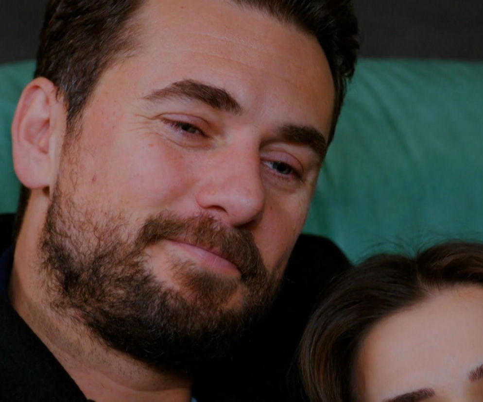 Uśmiechnięty Kuzey z brodą, w czarnym płaszczu i granatowej koszuli, spogląda w dół, tuląc do siebie Silę. Kobieta ma zamknięte oczy, długie, brązowe włosy. Oboje opierają się o zieloną kanapę. Więcej o ich wspólnej nocy w serialu Miłość i nadzieja na stronie Super Seriale.