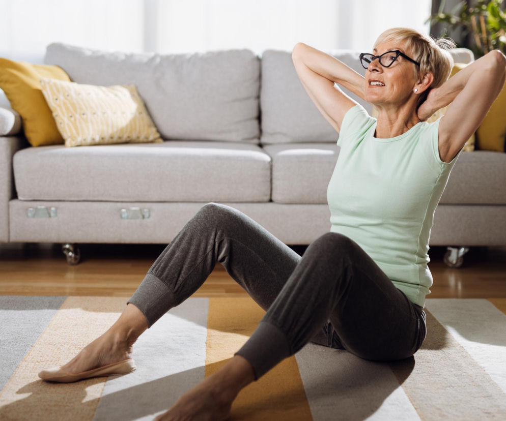 Uśmiechnięta, blondwłosa kobieta w okularach i jasnej koszulce wykonuje ćwiczenia na podłodze, z rękami za głową, siedząc w salonie z szarą kanapą w tle, co może pomóc w teście wstawania z podłogi, o którym przeczytasz na Poradnik Zdrowie.
