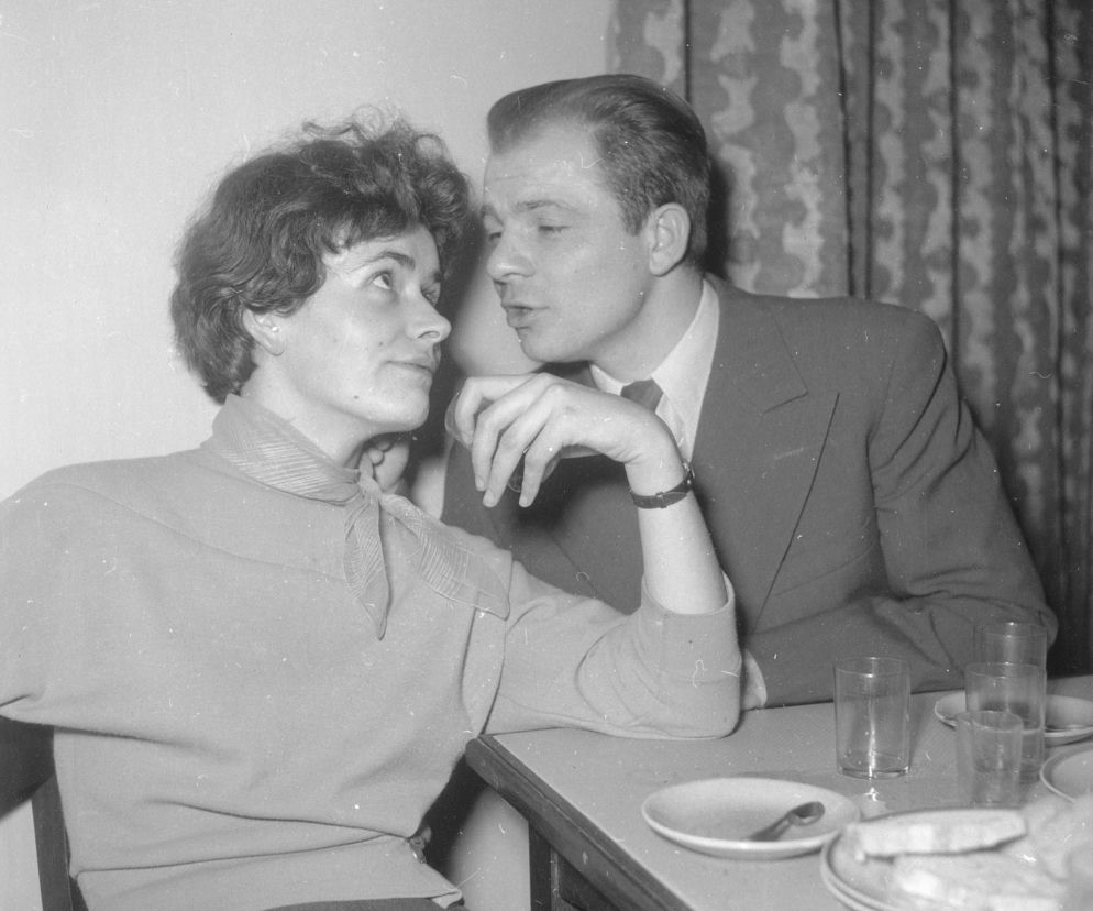 Młoda para, kobieta w jasnym swetrze z chustką na szyi i mężczyzna w garniturze, siedzi przy stole. On pochyla się, szepcząc jej coś do ucha, ona z zaciekawieniem na niego patrzy. Scena romantycznej randki w stylu PRL, nawiązująca do miłosnych hitów tamtych lat, o których przeczytasz na Super Biznes.