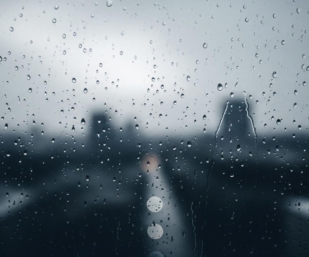 Szyba pokryta jest licznymi kroplami deszczu o różnej wielkości, rozrzuconymi na całej powierzchni. Tło za szybą jest rozmazane, przedstawiające zamglony, szary krajobraz miejski, z ciemnymi zarysami budynków. W centralnej części obrazu widoczne są rozmyte, jasne światła, tworzące efekt bokeh, przypominające latarnie lub inne źródła światła na ulicy, biegnącej od dołu do góry obrazu. Dominują odcienie szarości i błękitu, tworząc chłodną i melancholijną atmosferę.