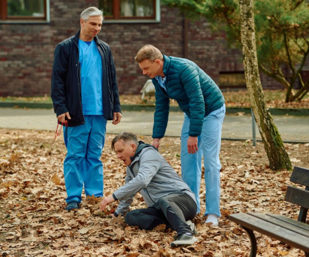 Greg Kalski i Kamil pomagają Adamskiemu wstać po wyczerpującym treningu. Tomasz leży na ziemi, a Greg i Kamil, obaj w odzieży medycznej, pochylają się nad nim, co widać w Na Wspólnej. Więcej o serialach na Super Seriale.
