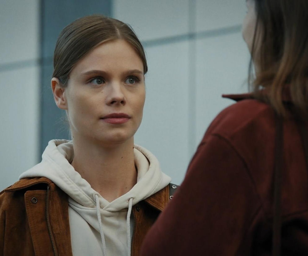 Kadr z serialu M jak miłość, przedstawiający Majkę (Matylda Giegżno) z jasnymi włosami, ubraną w brązową kurtkę i jasną bluzę z kapturem, rozmawiającą z inną postacią, której widać jedynie ramię. Majka zamyślona patrzy na rozmówczynię. Więcej na Super Seriale.