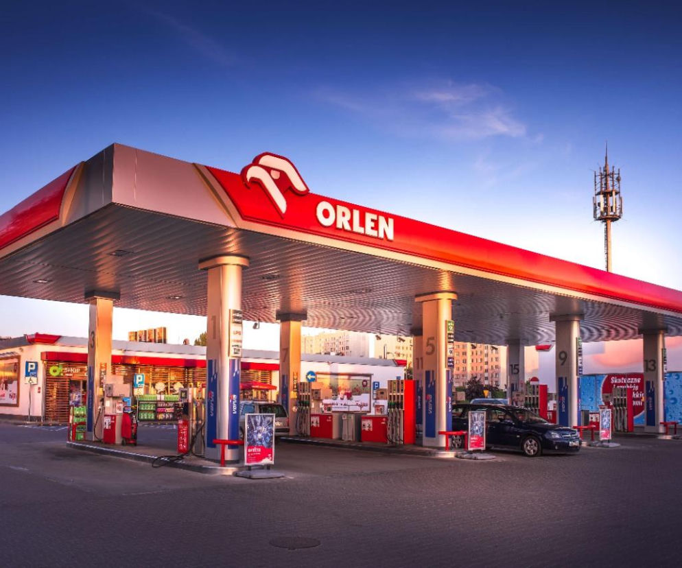 Stacja paliw Orlen z czerwonym daszkiem i białym logo Orlenu, zaparkowane samochody przy dystrybutorach. W tle widoczne budynki mieszkalne, słup telekomunikacyjny oraz inne punkty usługowe. W kontekście rekordowej dywidendy Orlenu, o której możesz przeczytać na Super Biznes.