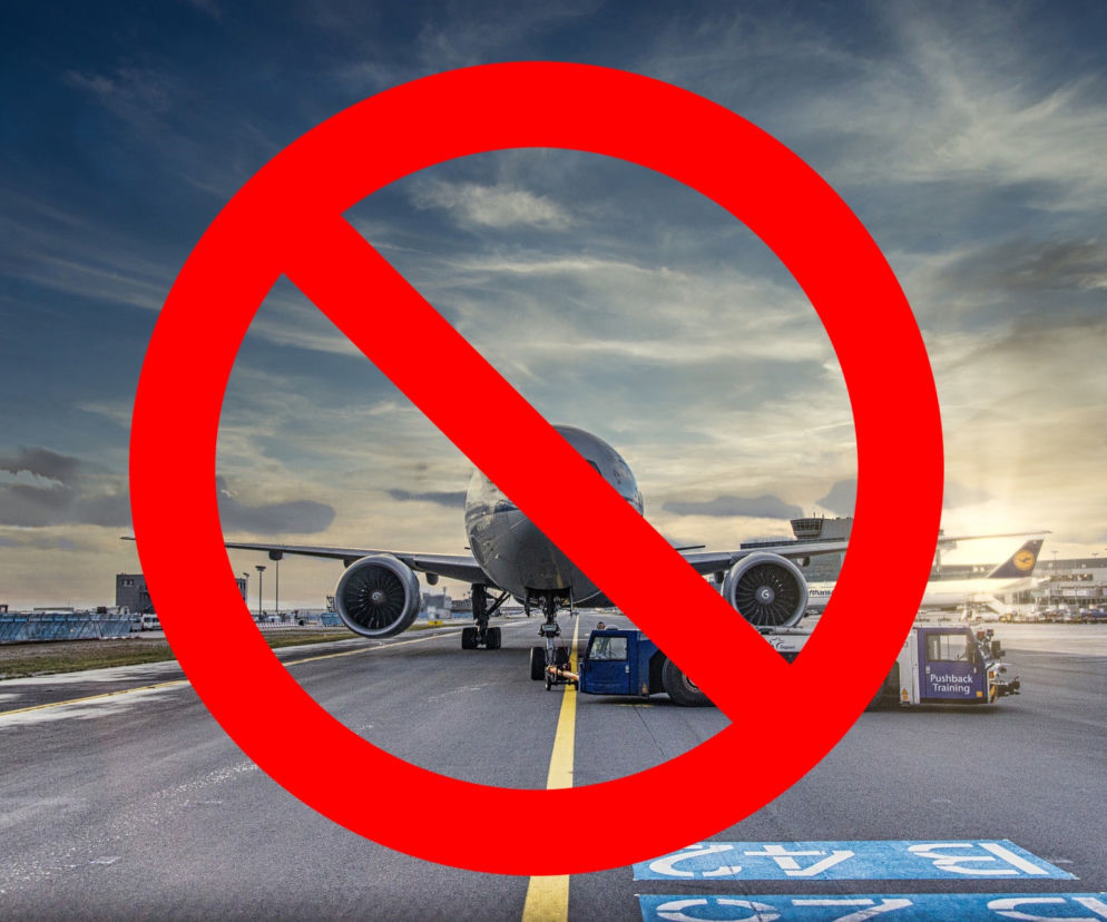 Duży samolot pasażerski na płycie lotniska z pojazdem do jego holowania, widoczny pod czerwonym okręgiem z przekreśleniem symbolizującym odwołane loty. Zachmurzone niebo o zachodzie słońca i światła lotniska w tle. Więcej informacji na Super Biznes.