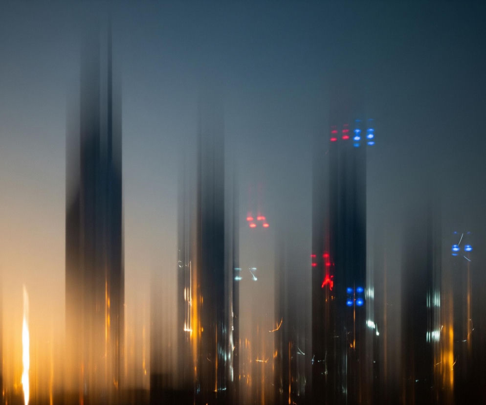 Abstrakcyjna kompozycja pionowych smug światła i ciemności dominuje w obrazie. U góry panuje ciemnoniebieski odcień, przechodzący w dół w szarość, a następnie w jasny pomarańczowo-złoty kolor, z najjaśniejszymi smugami światła na dole. Smugi są rozmyte i wydłużone, niektóre tworzą jasne, prawie białe pasma, podczas gdy inne są ciemniejsze, czarne lub granatowe, sugerując kształty budynków lub obiektów architektonicznych. W centralnej i prawej części obrazu widoczne są pojedyncze, rozmyte punkty świetlne w kolorach czerwonym i niebieskim, które również przyjmują wydłużone formy.