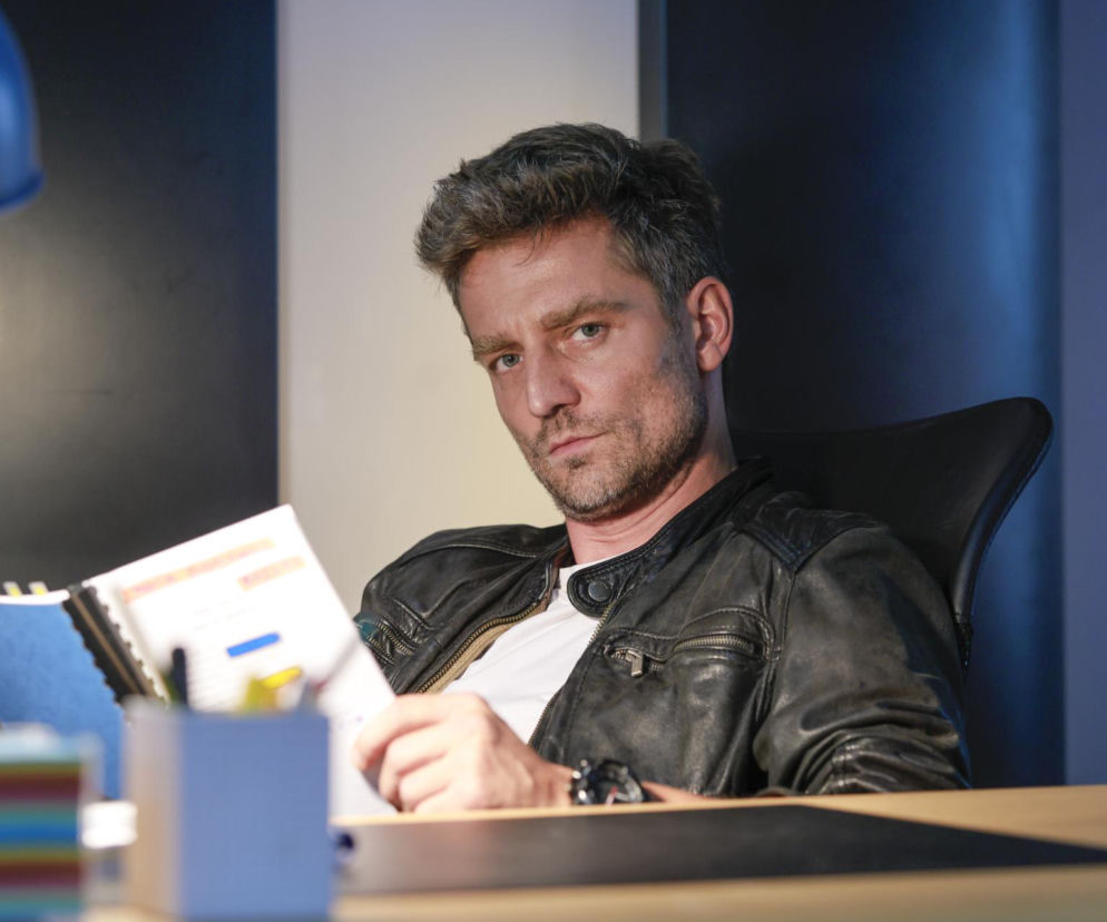 Aktor Marcin Rogacewicz w czarnej kurtce siedzi przy biurku z dokumentami. O jego sporze z byłą żoną przeczytasz na Eska.