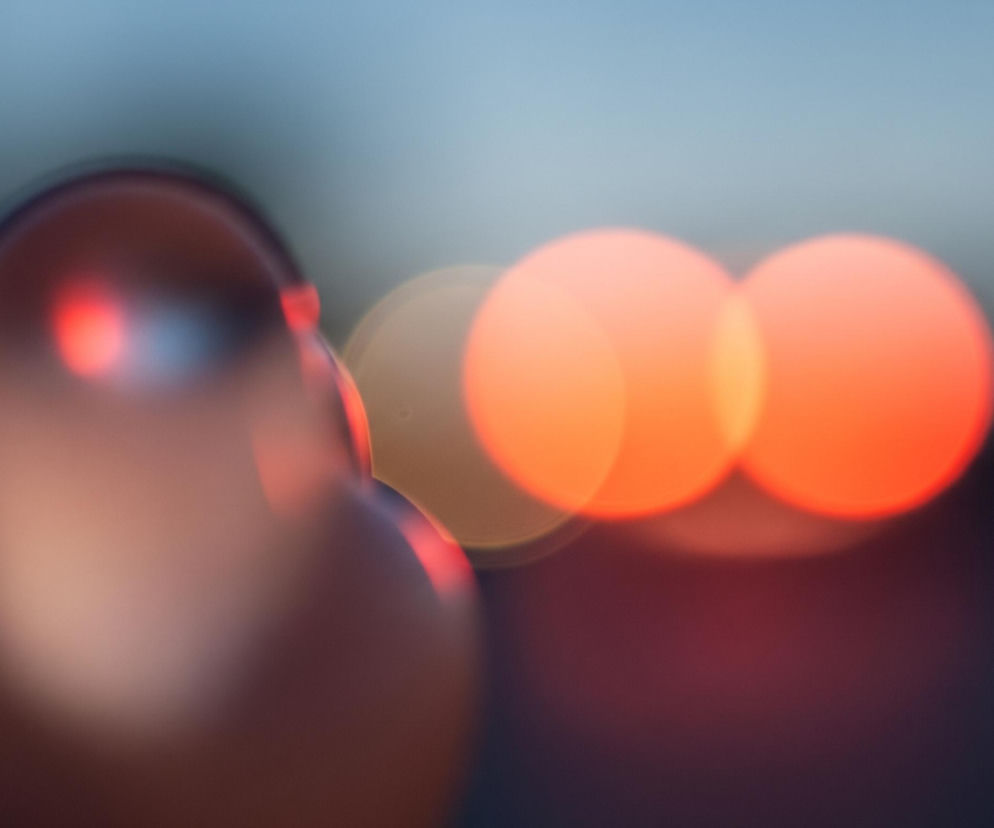Rozmyte, półprzezroczyste, zaokrąglone obiekty dominują lewą część kadru, mając odcień pomarańczowo-czerwony z jaśniejszymi smugami światła. Po prawej stronie widać dwa intensywnie pomarańczowe, okrągłe rozmyte punkty świetlne, przypominające światła tylne samochodu, umieszczone obok siebie na tle ciemnogranatowego, rozmytego nieba. Pod nimi znajduje się mniejszy, bardziej rozmyty czerwony punkt świetlny.