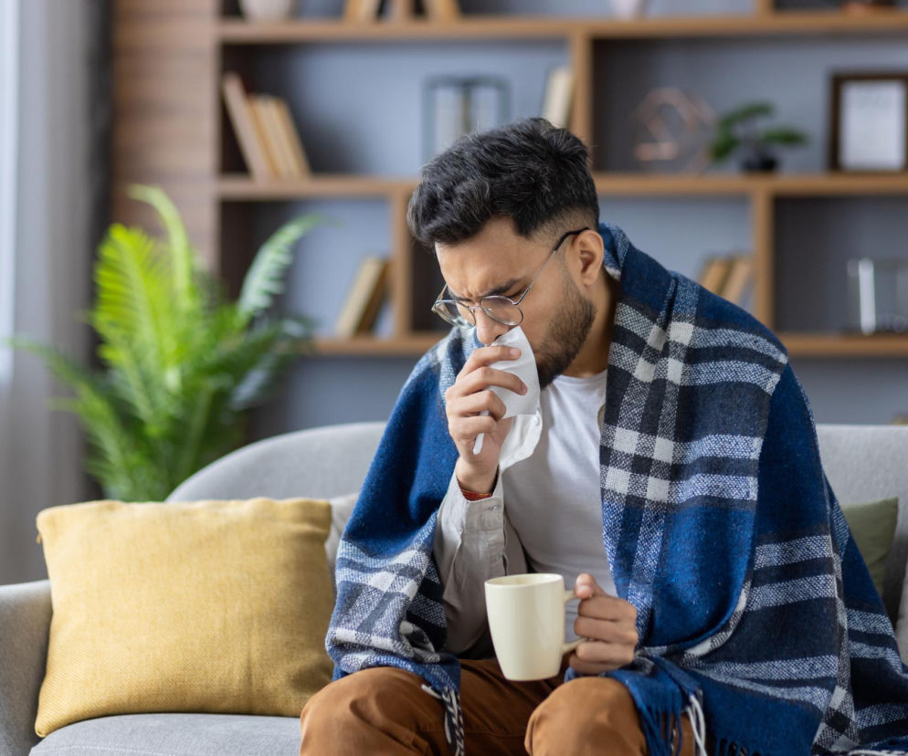 Mężczyzna z okularami, owinięty niebieskim kocem w kratę, siedzi na szarej kanapie, trzymając kubek i wycierając nos chusteczką, co symbolizuje objawy grypy w sezonie zachorowań, o których można przeczytać na portalu Poradnik Zdrowie.