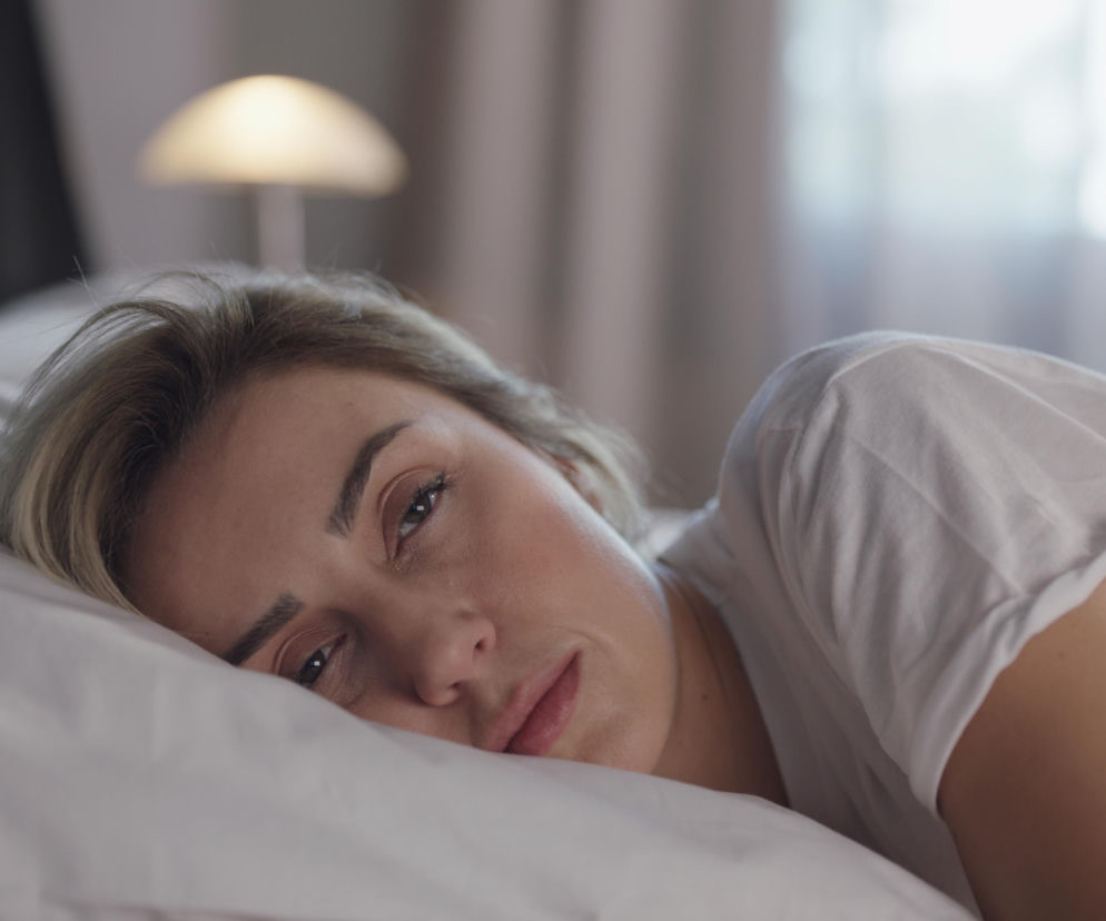 Kobieta z blond włosami leży na białej poduszce, ubrana w białą koszulkę, patrząc wprost na widza z lekko zmęczonym wyrazem twarzy. Zdjęcie ilustruje temat budzenia się przed budzikiem, o którym przeczytasz więcej na Poradnik Zdrowie.