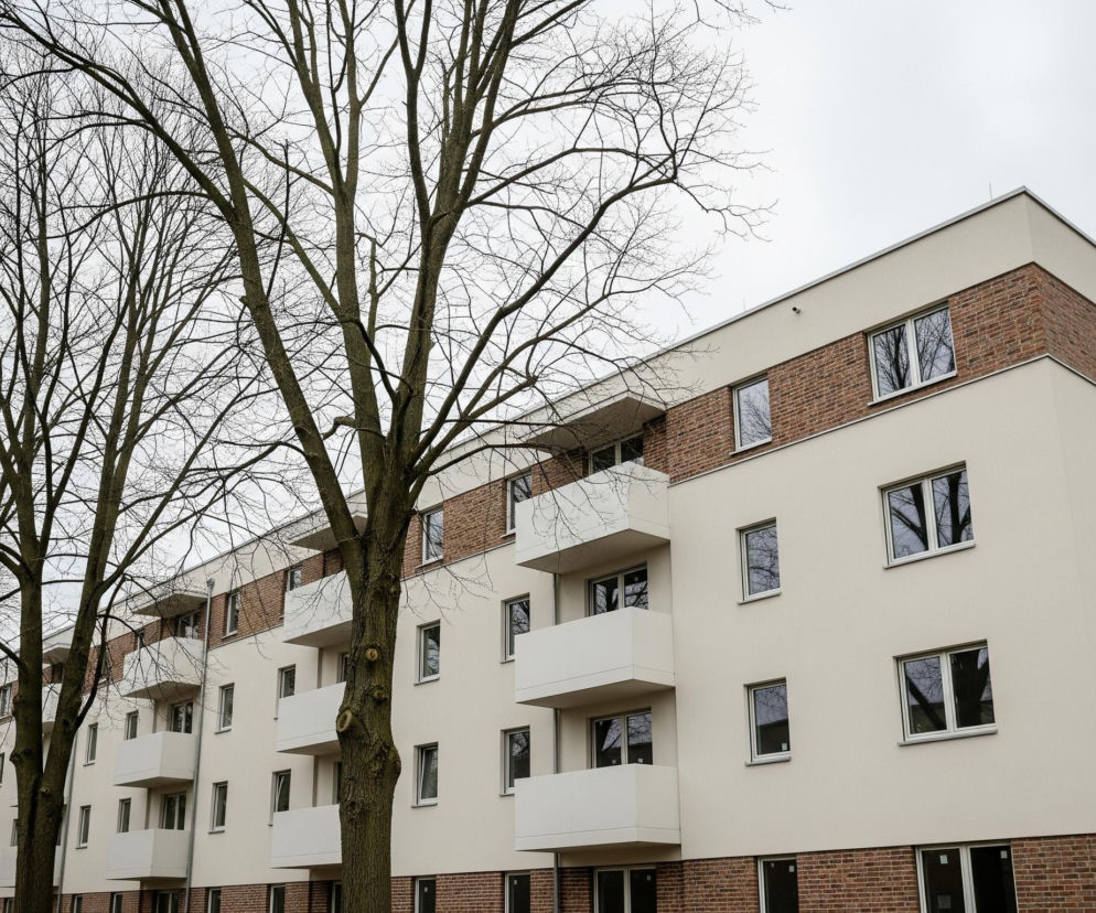 Duży, nowoczesny budynek mieszkalny o kremowych fasadach i fragmentach z czerwonej cegły dominuje prawą stronę kadru. Obiekt posiada szereg prostokątnych okien z białymi ramami oraz białe, kaskadowo ułożone balkony z balustradami. Przed budynkiem, po lewej stronie, rośnie rząd wysokich drzew liściastych o nagich, ciemnobrązowych gałęziach, które częściowo zasłaniają elewację. Niebo jest jednolicie jasnoszare, co sugeruje pochmurny dzień.