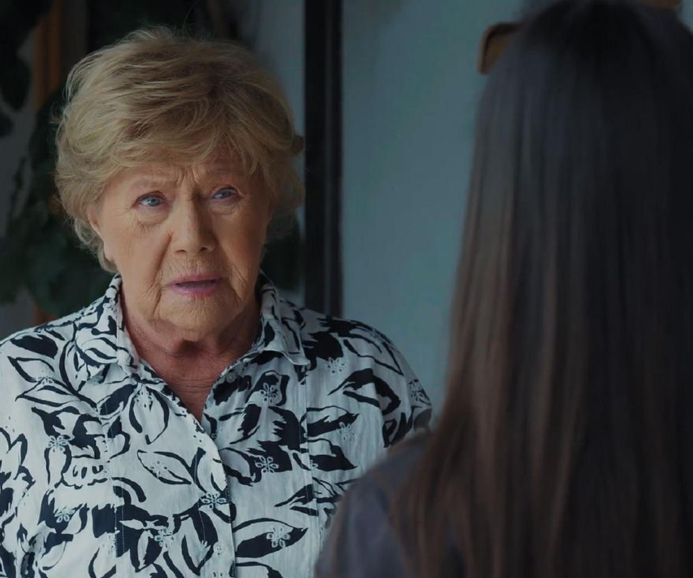 Barbara Mostowiak z serialu M jak miłość w kwiecistej koszuli, z krótkimi blond włosami, rozmawia z Basią, której tył głowy i długie ciemne włosy są widoczne na pierwszym planie. Kobiety poruszają trudną rodzinną tajemnicę, o której przeczytasz na Super Seriale.