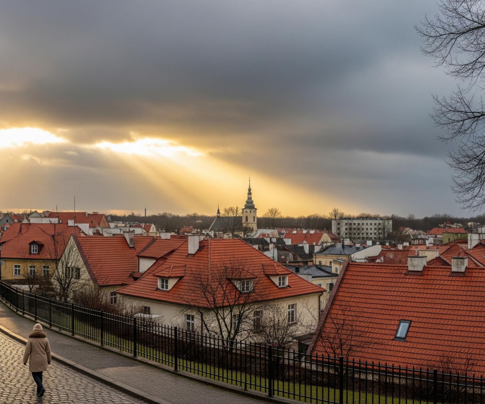 Kobieta w beżowej kurtce z futrzanym kołnierzem idzie brukowaną ulicą w dół, odwrócona tyłem do widza. Droga otoczona jest niskim, czarnym metalowym płotem z prawej strony i ciągiem kamienic z czerwonymi dachami. Horyzont domów zlewają się w tle z jasnym niebem rozświetlonym promieniami słońca przebijającymi się przez ciemne chmury. Po lewej i prawej stronie kadru widoczne są fragmenty nagich drzew.