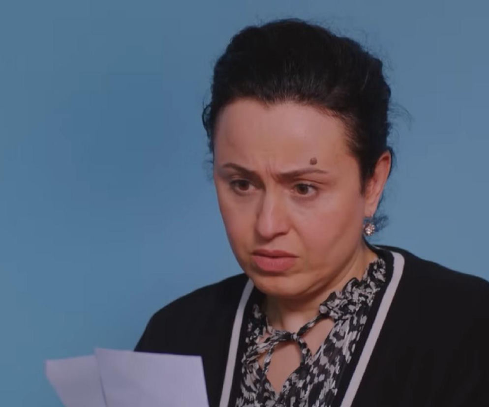 Kobieta o ciemnych włosach spiętych w kok, z widocznym pieprzykiem na czole, patrząca z niedowierzaniem na trzymane w rękach dokumenty. Ubrana jest w czarny rozpinany sweter na wzorzystej bluzce. Zdjęcie ilustruje emocje Cavidan z serialu Miłość i nadzieja w 363. odcinku. Więcej na Super Seriale.