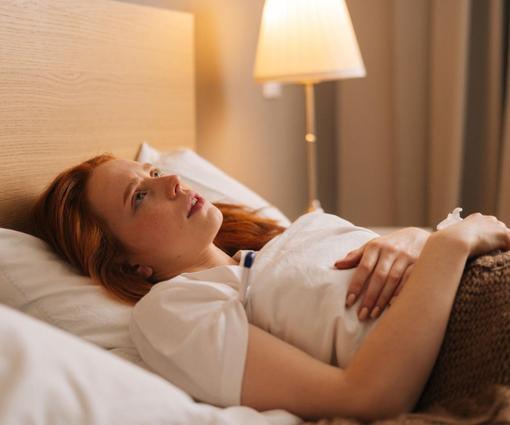 Kobieta o rudych włosach, z niepokojem leży w łóżku, trzymając się za brzuch, co symbolizuje problemy ze snem i trawieniem po późnej kolacji. Zdjęcie ilustruje negatywny wpływ wieczornego jedzenia na jakość snu, na temat którego więcej można przeczytać na portalu Poradnik Zdrowie.