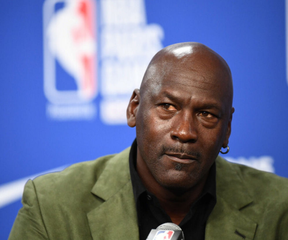 Koszykarz Michael Jordan w oliwkowej marynarce na tle logo ligi NBA. O aukcji jego koszulki przeczytasz na SE Supersport.