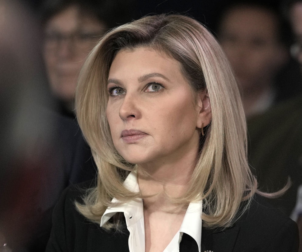 Blondwłosa Ołena Zełenska, Pierwsza Dama Ukrainy, w białej koszuli i czarnej marynarce, z wyrazem skupienia na twarzy. Tło jest rozmazane, z niewyraźnymi sylwetkami. Czytaj więcej o jej odwołanym wykładzie na naszym portalu.