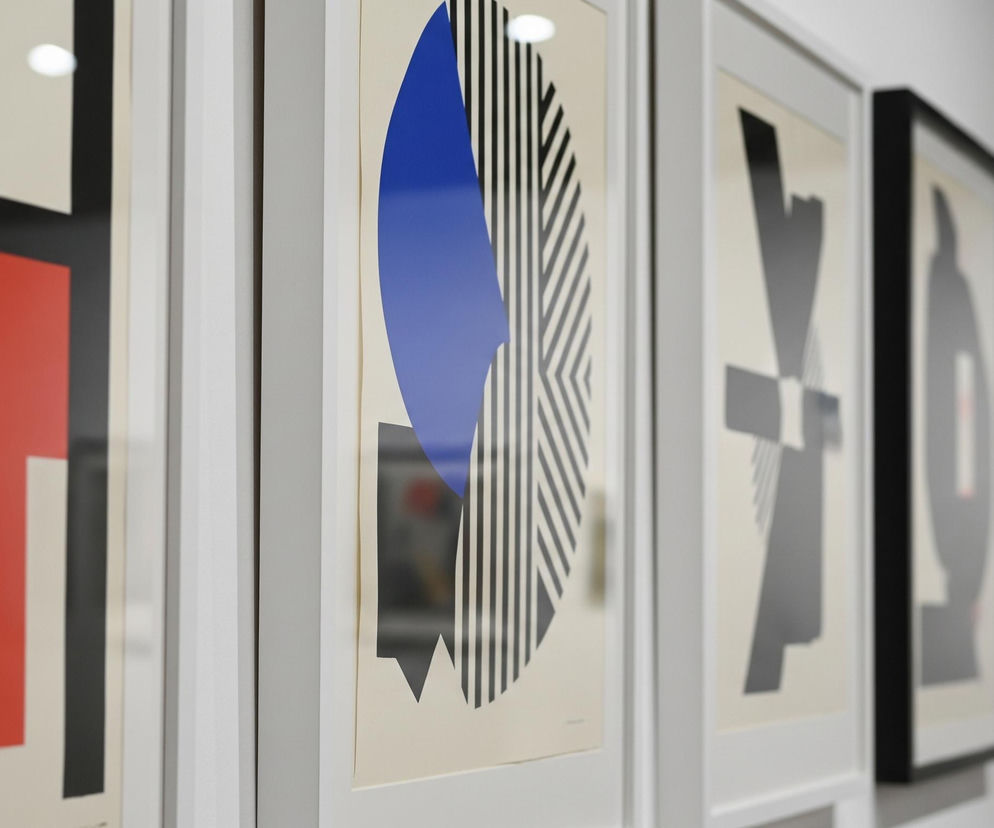 Ściana galerii sztuki eksponuje szereg oprawionych grafik o abstrakcyjnych wzorach geometrycznych. Na pierwszym planie, po lewej stronie, widoczny jest fragment obrazu w czerwono-czarno-kremowej kolorystyce, gdzie czerwone tło kontrastuje z czarnymi i kremowymi kształtami tworzącymi skomplikowaną kompozycję. Centralny obraz przedstawia dużą niebieską, owalną formę częściowo przesłoniętą czarno-białymi pasami, na kremowym tle, z wyraźnymi ostrymi zakończeniami kształtów. Obrazy, umieszczone w prostych białych ramach, odbijają światło z niewidocznych źródeł oświetlenia, tworząc jasne refleksy na ich powierzchni. Dalsze grafiki, poza ostrością, również utrzymane są w podobnej estetyce geometrycznych kształtów w kolorach czerni, szarości i kremu, a na końcu rzędu widać mniejszy, nieostry obraz z jaskrawymi czerwonymi elementami.