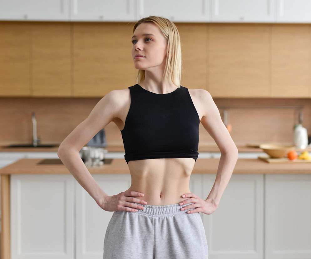 Młoda kobieta z syndromem klepsydry w kuchni, prezentująca napięcie mięśni brzucha, które powoduje charakterystyczne wklęśnięcie. Ta poza, omówiona na Poradnik Zdrowie, jest związana z bólem pleców, zgagą i problemami trawiennymi.