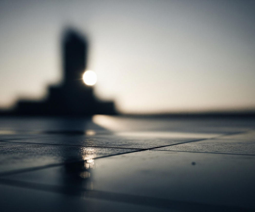 Pierwszy plan ukazuje ciemną, gładką powierzchnię podzieloną liniami na prostokątne płyty, na której odbijają się jasne punkty światła. Za nią, w oddali, znajduje się rozmyty kształt wysokiego budynku lub pomnika, który częściowo zasłania jaskrawe słońce, tworząc ciemną sylwetkę na tle jasnego horyzontu. Tło to rozległe, bezchmurne niebo, przechodzące od jasnoszarego na dole do ciemnoszarego u góry, z wyraźnym jasnym punktem słońca po prawej stronie centralnej części kadru. Całość utrzymana jest w chłodnej tonacji szarości i błękitu, z mocnym kontrastem między rozjaśnionym słońcem a ciemnymi elementami na pierwszym planie i w tle.