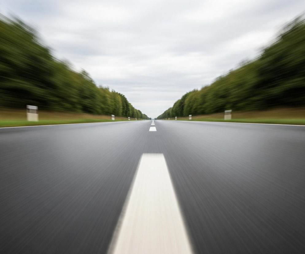 Asfaltowa droga rozciąga się w głąb obrazu, z wyraźną, białą linią ciągłą na środku, która staje się cieńsza i jaśniejsza w oddali. Po obu stronach drogi widoczne są rozmazane, zielone pasy drzew, tworzące wrażenie szybkiego ruchu. Trawa o brązowo-zielonym odcieniu leży u podstawy drzew, oddzielając je od szarej nawierzchni drogi. Szare, zachmurzone niebo zajmuje górną część kadru, również rozmazane, co potęguje dynamiczny efekt.