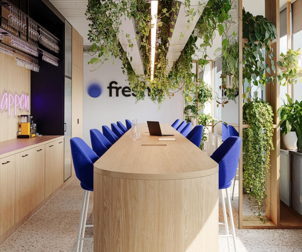 Nowoczesne biuro Fresha w Warszawie, projektu SAAN architekci, prezentuje długi, drewniany stół w fioletowej strefie socjalnej. Obok wiszą lampy z bujną zielenią. Można o tym przeczytać na portalu Architektura Murator Plus.