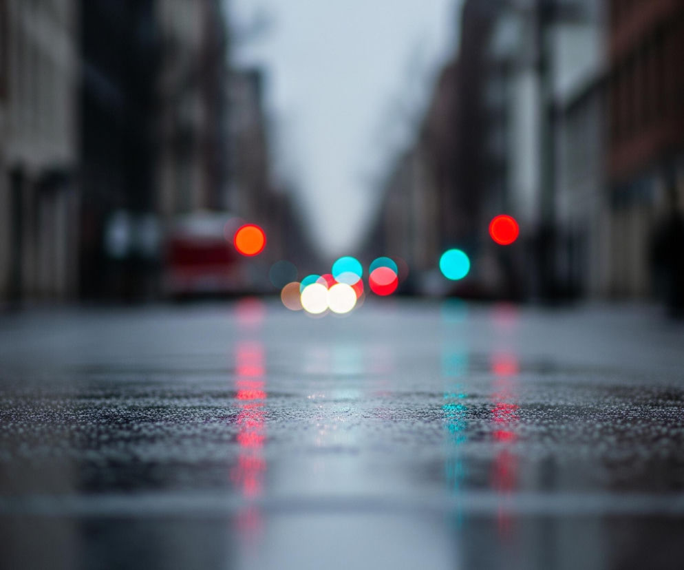 Mokry asfaltowy pas drogi wypełnia dolną połowę obrazu, z mnóstwem drobnych kropli wody widocznych na jego powierzchni, odbijających rozmazane światła. W oddali, w górnej połowie kadru, widać szereg kolorowych, okrągłych świateł bokeh: czerwone, pomarańczowe, białe i turkusowe, rozmieszczone wzdłuż ulicy. Po obu stronach drogi, w tle, zarysowują się niewyraźne, ciemne sylwetki budynków, wtapiające się w szarawe, zamglone niebo.