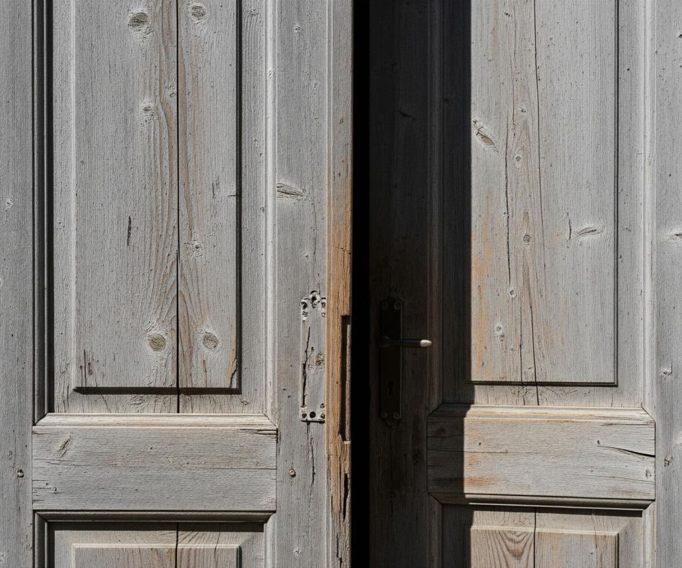 Pionowo ujęte, lekko uchylone dwuskrzydłowe drewniane drzwi w odcieniach szarości i brązu, flankowane po bokach przez jasną, teksturowaną ścianę. Lewe skrzydło drzwi jest nieco bardziej zamknięte niż prawe, a pomiędzy nimi widoczna jest ciemna szczelina. Na obu skrzydłach widoczne są sęki i spękania drewna, a także metalowe okucia i zawiasy, z czego pośrodku uchylonych drzwi widoczny jest metalowy element zamka oraz klamka.