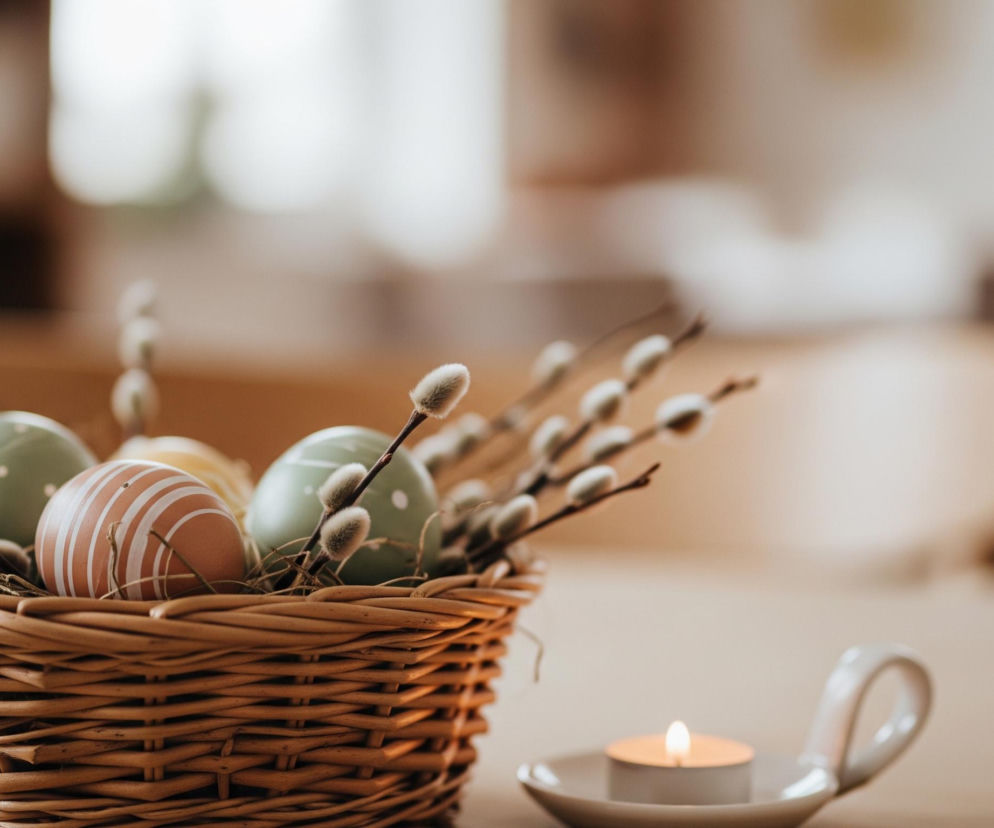 Wiklinowy koszyk z pisankami i gałązkami bazi stoi na jasnym, drewnianym stole, zajmując centralną część kadru po lewej stronie. W koszyku znajdują się różnokolorowe jajka, w tym jedno brzoskwiniowe z białymi paskami, jedno miętowe z białymi kropkami oraz częściowo widoczne żółte i inne miętowe jajka. Pomiędzy pisankami widać puszyste bazie w odcieniach szarości i brązu, wystające ponad krawędź koszyka. Na prawo od koszyka, bliżej dolnej krawędzi, znajduje się mała świeczka typu tealight płonąca w białym świeczniku z uchwytem. Tło jest rozmyte, ukazując jasne, ciepłe barwy i niewyraźne kształty przedmiotów, co sugeruje wnętrze pomieszczenia.