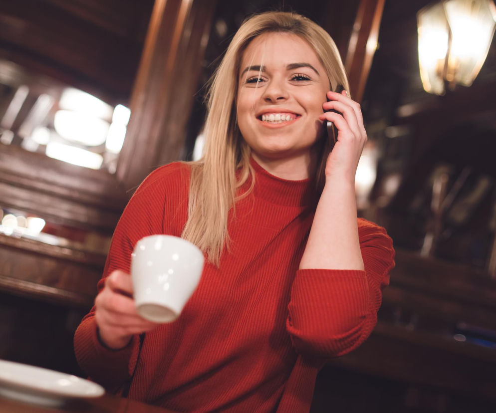 Uśmiechnięta blondynka w czerwonym swetrze trzyma filiżankę kawy i telefon przy uchu, siedząc w kawiarni, co symbolizuje codzienny rytuał picia kawy. Dowiedz się więcej o związku kawy z ryzykiem problemów z nerkami na Poradnik Zdrowie.