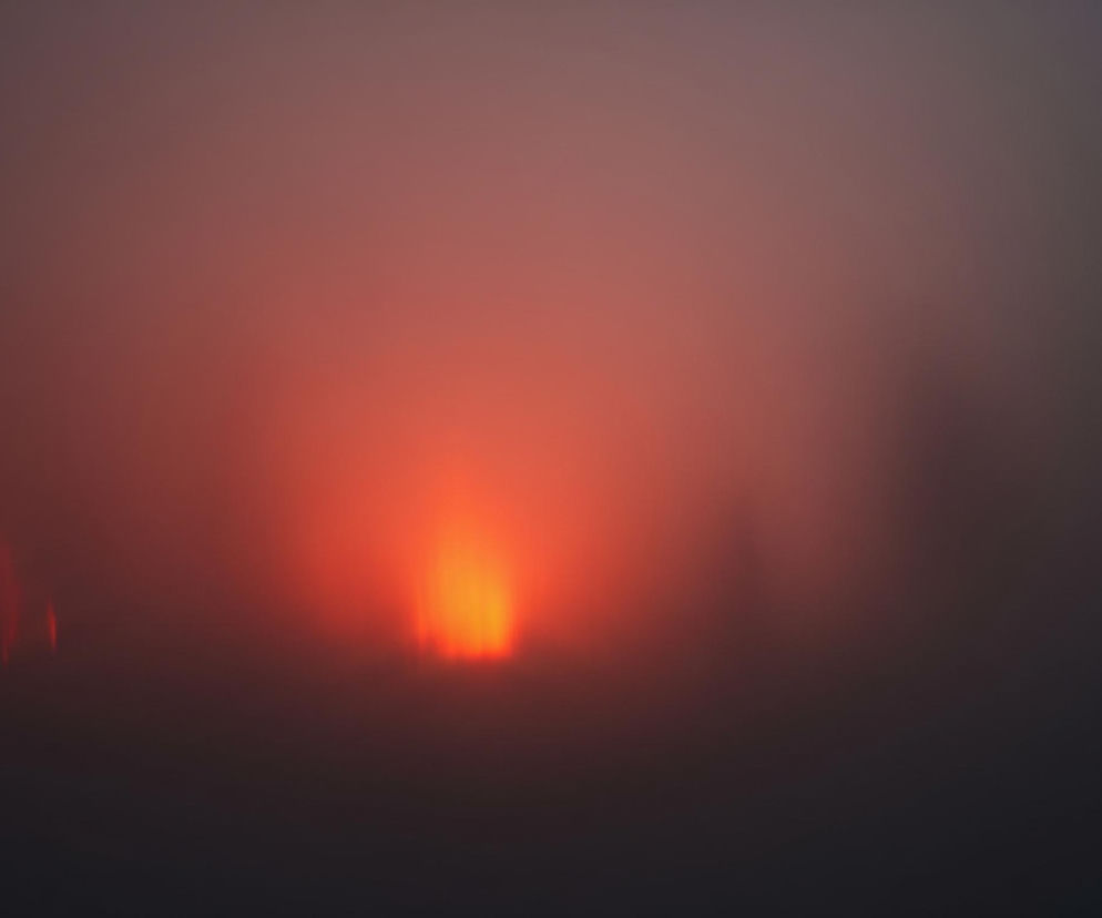 Centralnie na obrazie znajduje się jaskrawy, owalny kształt w odcieniach intensywnego pomarańczu i czerwieni, który rozświetla otaczającą go mglistą przestrzeń. Światło to stopniowo przechodzi w ciemniejsze, rozmyte barwy brązu i szarości, dominujące w górnej i dolnej części kadru. Po lewej stronie, w tle, widoczne są trzy pionowe, rozmyte czerwone smugi. Całość kompozycji jest zamazana i brakuje jej ostrości.