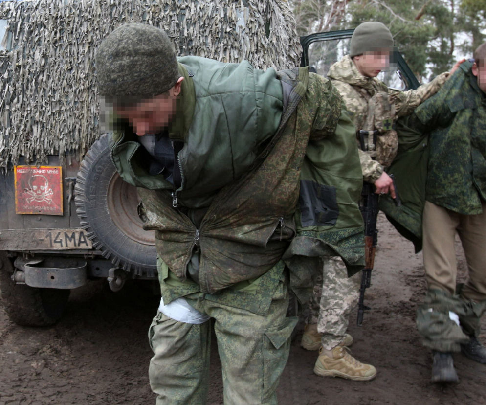 Trzech jeńców wojennych w wojskowych ubraniach, z pikselizowanymi twarzami, stoi przy zamaskowanym samochodzie. Jeden Ukrainiec eskortuje Rosjanina. O ich losach przeczytasz na Super Biznes.